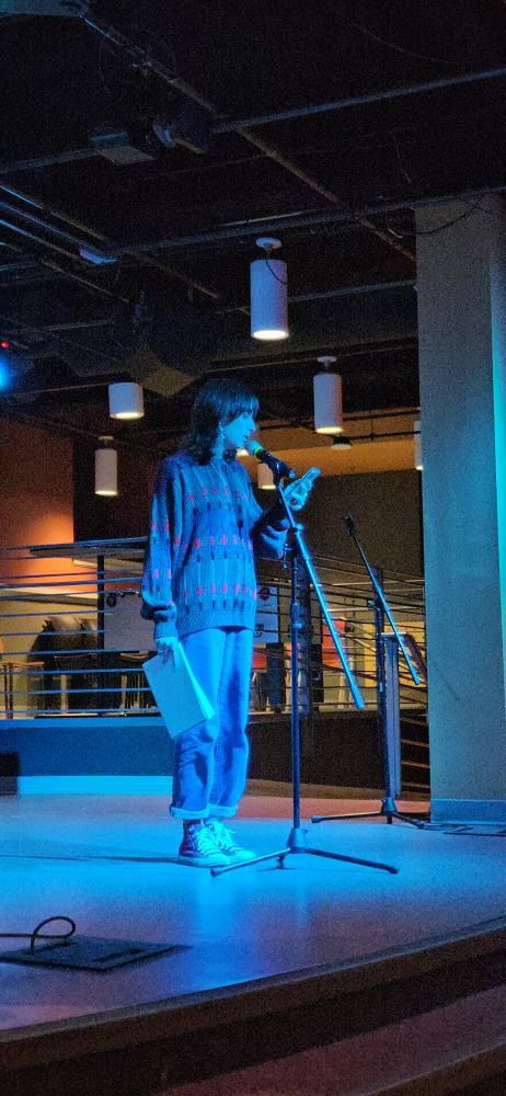 A woman in blue stands in front of a microphone on a stage, reading something from her phone