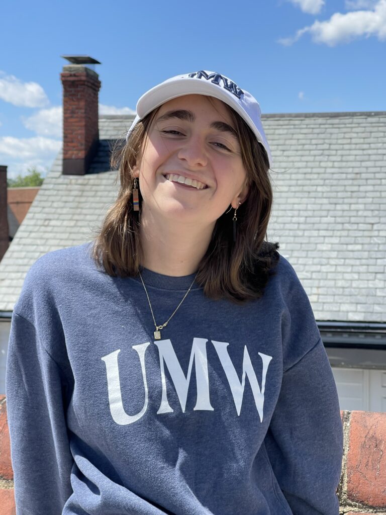 A woman wearing a white hat and a blue "UMW" sweatshirt smiles at the camera. 