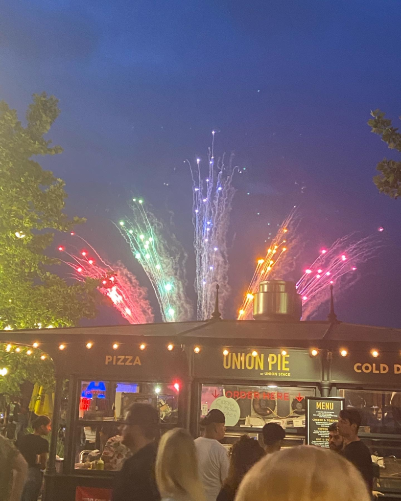 Rainbow colored fireworks explode over a pizza stand that reads "PIZZA- UNION PIE- COLD DRINKS"